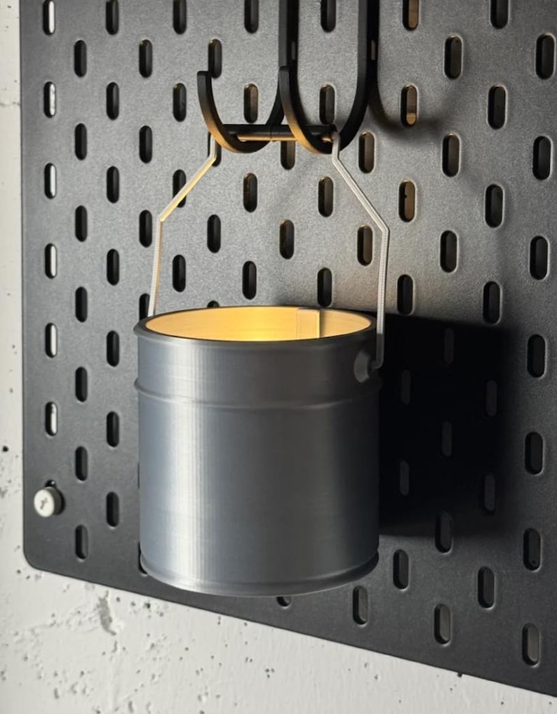 May include: A gray metal bucket-shaped candle holder suspended from a black pegboard. The candle holder has a cylindrical shape with a light source inside, casting a warm glow. The pegboard has evenly spaced oval holes.