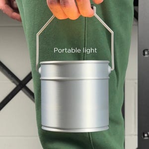 May include: A silver-colored portable light with a handle. The cylindrical light has a handle made of thin metal. The text "Portable light" is printed on the front of the light.