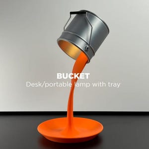 May include: A distinctive desk lamp featuring a silver bucket-shaped shade, seemingly pouring orange light onto a matching tray. The lamp has a black handle and the text "BUCKET Desk/portable lamp with tray".
