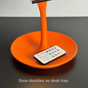 May include: An orange desk lamp with a circular base that doubles as a tray. The lamp has a slender stem and a silver support at the top. A remote control with multiple colored buttons rests on the tray. The text "Base doubles as desk tray" is visible.