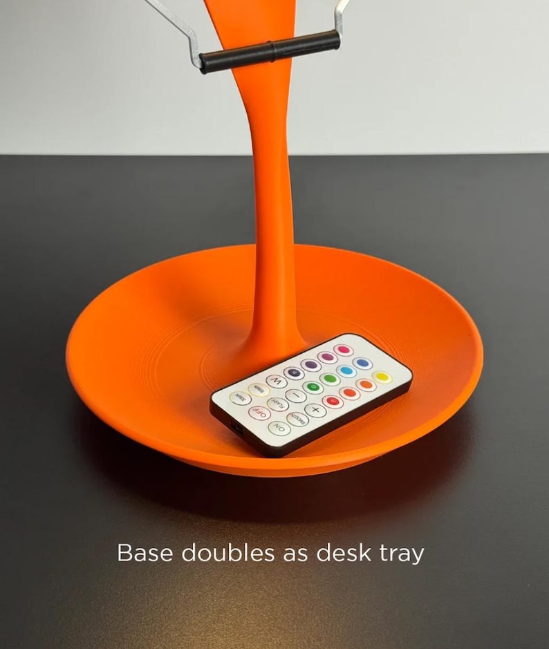 May include: An orange desk lamp with a circular base that doubles as a tray. The lamp has a slender stem and a silver support at the top. A remote control with multiple colored buttons rests on the tray. The text "Base doubles as desk tray" is visible.