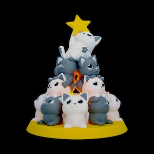 May include: A whimsical cat figurine arrangement. The sculpture features a pyramid of gray, white, and pink cat figures, topped with a yellow star. The base is a yellow circle. The cats have large eyes and are in various poses.