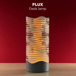 May include: A cylindrical desk lamp labeled FLUX with a solid dark gray base and a series of stacked, white, wavy horizontal layers that emit a warm orange glow from the center. The lamp features a distinct undulating texture throughout its body.