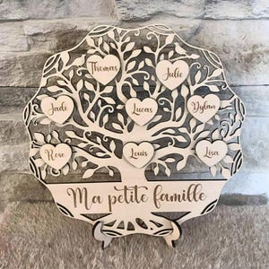 Peut inclure: Un arbre généalogique en bois avec des étiquettes en forme de cœur portant des noms. L'arbre est orné de feuilles et de branches découpées, avec les noms Thomas, Julie, Jade, Lucas, Dylan, Rose, Louis et Lisa. La phrase "Ma petite famille" est en bas.