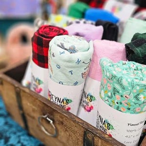 Op de afbeelding: Een houten kist gevuld met opgerolde vierkante stukken stof. De stofvierkanten zijn in verschillende patronen, waaronder ruit, bloemen en alfabetprints. De stofvierkanten zijn gelabeld met de tekst "REYNA'S SEWING".