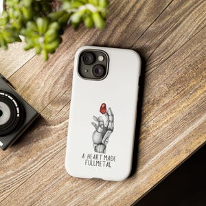 May include: White iPhone case featuring a black and white illustration of a robotic hand holding a red heart. The text "A HEART MADE FULLMETAL" is printed below the illustration.