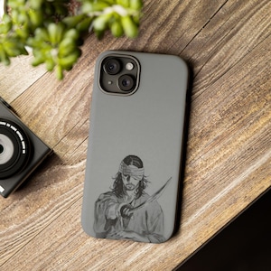 May include: A gray phone case with a black and white illustration of a blindfolded person holding a sword.