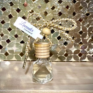 May include: A small glass bottle with a wooden cap and a gold and white string tied around the neck. The bottle is filled with a clear liquid and has a label that says "Lavender Essential Oil".