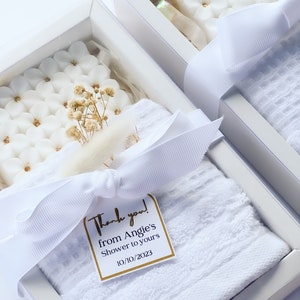 May include: White gift box with a white towel, a white soap bar with a floral design, and dried flowers. The box is tied with a white ribbon and has a gold tag that says "Thank you! from Angie's Shower to yours 10/10/2023".