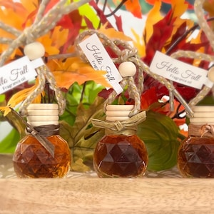 Op de afbeelding: Drie kleine glazen flesjes met bruine vloeistof, elk met een kurkstop, een touw strik en een label met de tekst "Hello Fall Fragrance Oil". De flesjes staan op een houten oppervlak met een achtergrond van herfstbladeren.