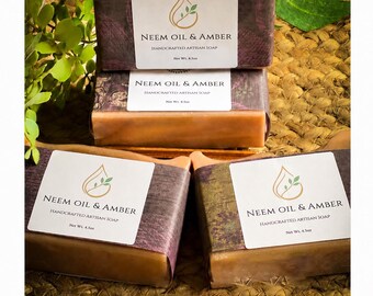 May include: Four rectangular bars of orange-toned soap stacked and arranged, each wrapped in a dark purple paper band with a white label reading Neem Oil & Amber Handcrafted Artisan Soap Net Wt. 4.5oz, featuring a gold droplet logo with a green leaf.