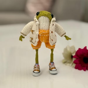 May include: A green knitted frog wearing a white cardigan, orange shorts, and white sneakers. The frog is standing on a white surface.
