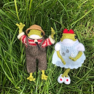 May include: Two crocheted frog plushies, one wearing a brown hat, red and white striped sweater, and brown overalls, the other wearing a blue dress with white stars and red shoes.