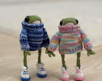May include: Two knitted frog plushies wearing sweaters and shoes. One frog is wearing a blue and white striped sweater and blue shoes. The other frog is wearing a pink, yellow, and white striped sweater and pink shoes.
