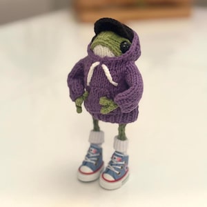 May include: A handmade, knitted frog figurine wearing a purple hoodie, black cap, and blue sneakers. The frog has green skin, white socks, and a white drawstring on the hoodie. The sneakers have white soles and red accents.