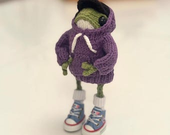 May include: A handmade, knitted frog figurine wearing a purple hoodie, black cap, and blue sneakers. The frog has green skin, white socks, and a white drawstring on the hoodie. The sneakers have white soles and red accents.