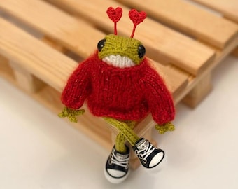 May include: A green knitted frog wearing a red sweater and black sneakers with red hearts on its head. The frog is sitting on a wooden bench.