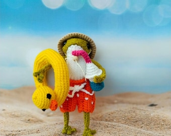 Puede incluir: Una rana de crochet con un sombrero de paja, pantalones cortos naranjas y un flotador inflable amarillo. La rana sostiene una nevera de crochet azul y blanca. La rana está de pie en una playa de arena.