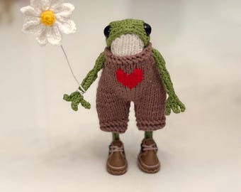 May include: A small, green, crocheted frog figurine wearing brown knit overalls with a red heart patch on the chest. The frog has a white face, black eyes, and is holding a white crocheted daisy flower with a yellow center. It wears brown shoes.