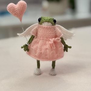 Puede incluir: Figura de rana tejida a mano, vestida con un vestido rosa, alas de ángel blancas y zapatos blancos. La rana sostiene un globo en forma de corazón rosa. La rana tiene una cabeza verde y ojos negros. El fondo está borroso.