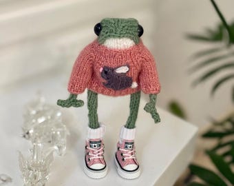 May include: A green knitted frog wearing a pink sweater with a brown bunny applique and pink Converse sneakers.