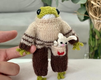 May include: A green knitted frog wearing a brown and beige striped sweater and brown pants. The frog is holding a small white knitted bunny in its pocket.