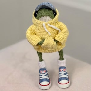 Puede incluir: Una rana de punto que lleva una sudadera amarilla con cordón blanco, una gorra de béisbol azul y blanca y zapatillas azules con suela blanca.