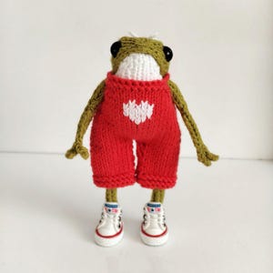 May include: A green knitted frog wearing red overalls with a white heart design and white sneakers.
