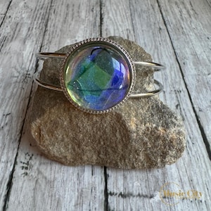 May include: A silver cuff bracelet with a large iridescent round cabochon. The cabochon has a green, blue, and purple color scheme.