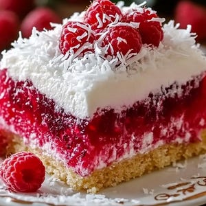 Puede incluir: Una porción de postre con una base dorada, una capa de gelatina roja vibrante y una gruesa cobertura de crema blanca espolvoreada con coco rallado. Las frambuesas rojas frescas coronan el postre, presentado en un plato decorativo.