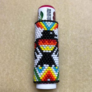 Puede incluir: Un porta bálsamo labial de cuentas blancas, negras, rojas, naranjas, amarillas, azules y verdes con un patrón geométrico. El tubo de bálsamo labial es blanco con las palabras "SEALED FOR PROTECTION" impresas en él.