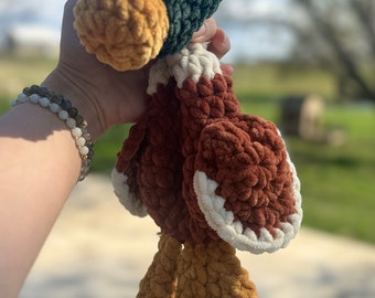 May include: A crocheted plush toy duck with a green head, brown body, and white accents. The duck is held in a hand with a bracelet.