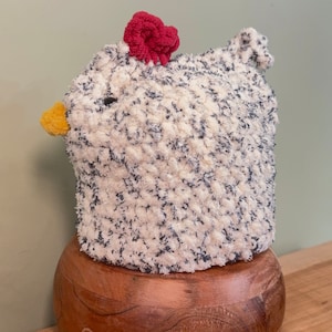 Könnte beinhalten: Ein weißes und schwarzes Häkel-Hühner-Spielzeug mit einem roten Bommel auf dem Kopf. Das Huhn sitzt auf einem Holzsockel.