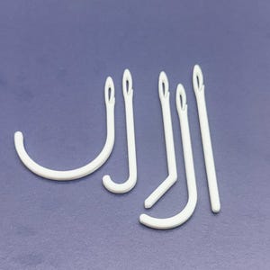 May include: A collection of five white, J-shaped hooks. The hooks are made of a smooth material and have a small hole at the top. They are arranged on a solid, blue-grey background.