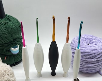 May include: A collection of crochet hooks with various colored metal hooks and white, black, and white handles. A green ball of yarn and a purple ball of yarn are in the background. The crochet hooks are for crafting and knitting.