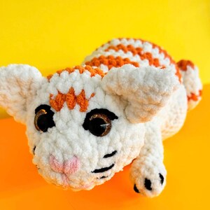 May include: A white crocheted cat toy with orange stripes and black eyes. The cat is lying on its side with its head turned to the side.