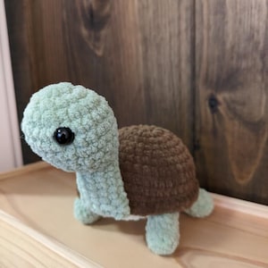 May include: A crocheted stuffed turtle toy with a brown shell and green head and legs. The toy has large black eyes and a fuzzy texture.
