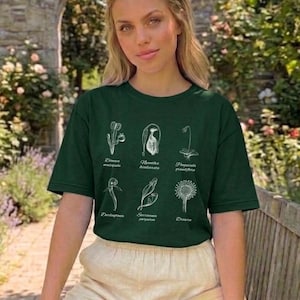 Puede incluir: Una camiseta verde oscuro con ilustraciones botánicas blancas y texto. Se combina con pantalones cortos beige. La imagen también incluye un libro y gafas de sol. El logotipo de The Garrison Trend es visible.