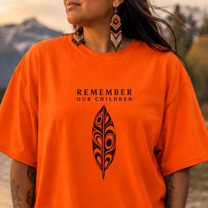 Op de afbeelding: Een feloranje T-shirt met korte mouwen voorzien van de zwarte tekst REMEMBER OUR CHILDREN boven een zwarte afbeelding van een gestileerde veer. Het ontwerp is gecentreerd op de borst van het kledingstuk, dat een ronde hals en een ruime pasvorm heeft.