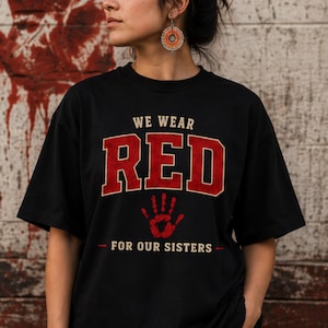 May include: A black short-sleeved t-shirt featuring the text WE WEAR RED FOR OUR SISTERS in a collegiate font. The word RED is in large red letters, and a red handprint graphic is centered below the text on the front of the garment.
