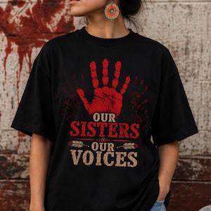 May include: A black short-sleeved t-shirt featuring a red handprint graphic on the chest. Below the handprint, the text reads "OUR SISTERS" in red and "OUR VOICES" in a textured tan color, separated by two horizontal feather illustrations.