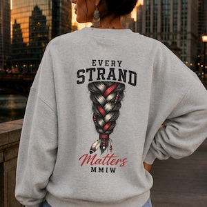 May include: A light gray crewneck sweatshirt featuring a graphic on the back of a dark braided hairstyle with red accents and a feather. The text reads EVERY STRAND Matters MMIW in black and red lettering. The sweatshirt has long sleeves.