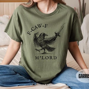 May include: Olive green t-shirt featuring a black crow illustration holding a sword, with the text "F-CAW-F M'LORD". The shirt is a comfortable fit and made of soft material.