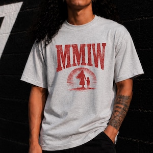 May include: A light gray short-sleeved t-shirt featuring a distressed red graphic print. The design includes the text MMIW in a bold arched font above a silhouette of an adult and a child standing together against a circular sun-like background.