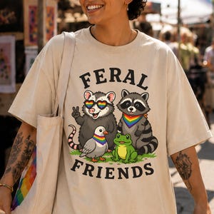 Peut inclure: Un t-shirt beige avec une illustration représentant un opossum, un raton laveur, un pigeon et une grenouille. L'opossum porte des lunettes de soleil arc-en-ciel en forme de cœur. Le texte FERAL FRIENDS est imprimé en lettres noires.