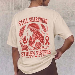 Può includere: Una t-shirt a maniche corte color crema con una grafica rossa sul retro. Il design include il testo "STILL SEARCHING" e "STOLEN SISTERS" che circonda l'illustrazione di una persona, un'impronta di mano, piume, rose e un nastro.