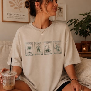 Puede incluir: Camiseta beige con cuatro ilustraciones botánicas y textos: Sundew, Venus Flytrap, Butterwort y Nepenthes. La camiseta es de corte holgado y tiene mangas cortas. El logotipo de The Garrison Trend es visible en la esquina inferior derecha.
