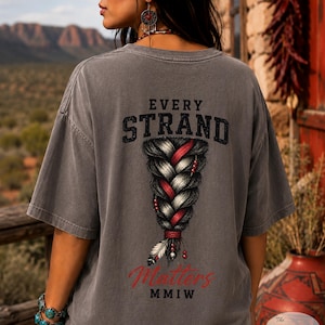 May include: A charcoal gray short-sleeve t-shirt featuring a graphic on the back of a thick, braided hairstyle with red and white highlights. The text reads EVERY STRAND Matters MMIW, with a small feather accent at the base of the braid.