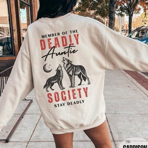 May include: Cream-colored sweatshirt with the text "MEMBER OF THE DEADLY Auntie SOCIETY STAY DEADLY" in red and black. The graphic features two wolves howling at a crescent moon. The Garrison Trend logo is in the bottom right corner.