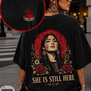 May include: A black t-shirt featuring a graphic on the back of a person with red handprints on their cheeks, surrounded by red roses and foliage, with the text "SHE IS STILL HERE". The front chest area displays a small red handprint and flowers.